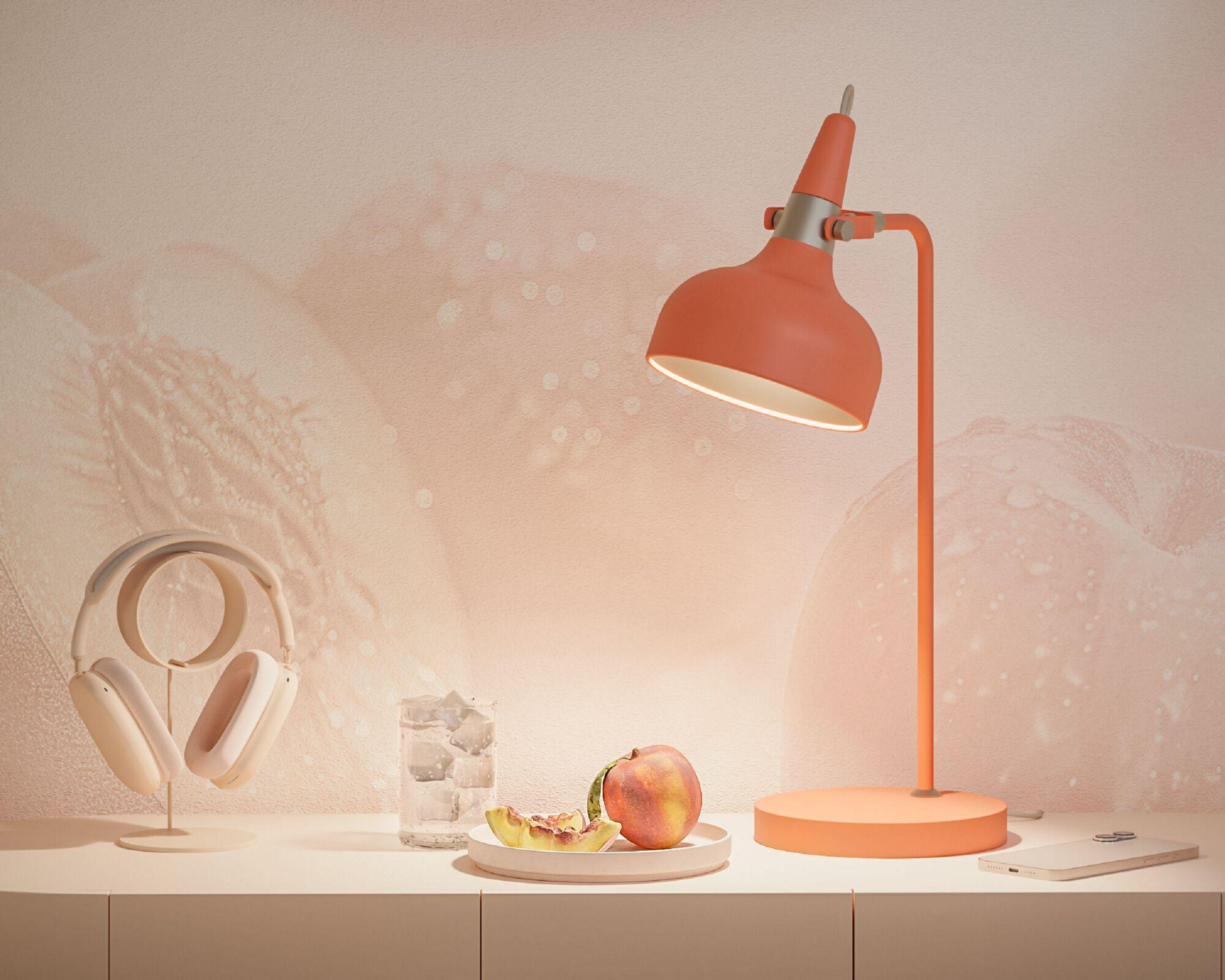 Lampa biurkowa w kolorze brzoskwini, słuchawki, plasterki brzoskwini i brzoskwinia na talerzu. Tło jest brzoskwiniowe.