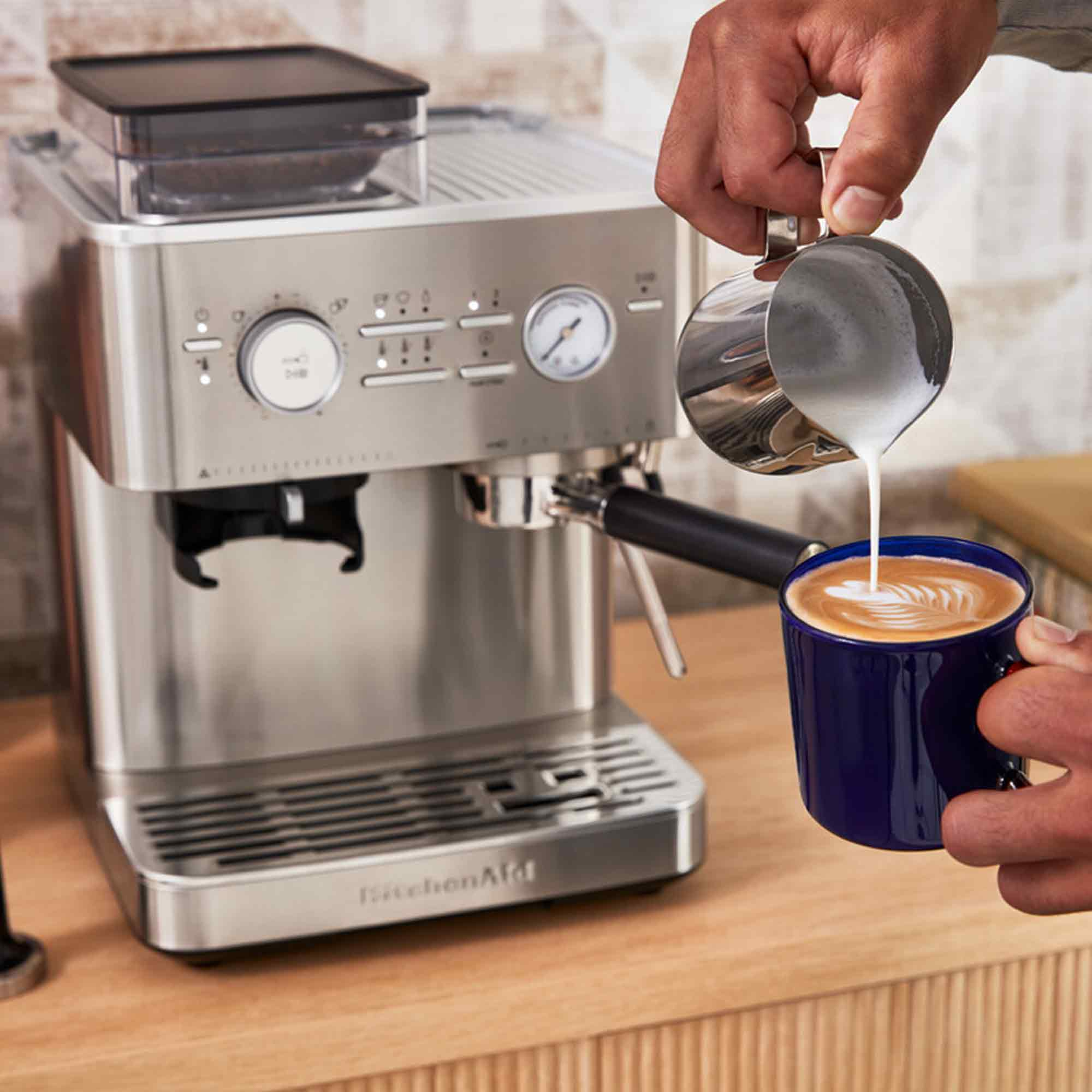Eine Person gießt Milch in einen blauen Kaffeebecher. Eine Espressomaschine steht auf Holz.