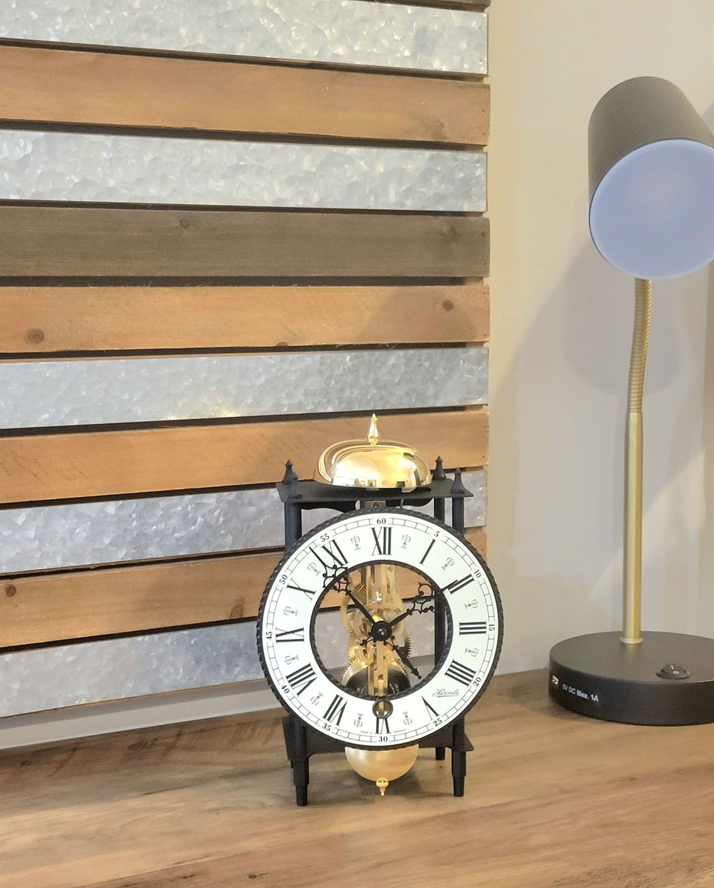 Eine Uhr mit römischen Ziffern steht auf einer Holzoberfläche. Rechts ist eine Lampe. Der Hintergrund ist eine Holz- und Metallwand.