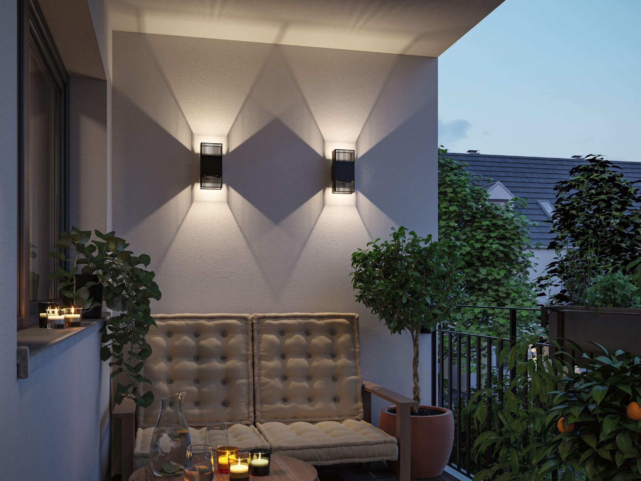 Scène de balcon avec appliques murales, sièges, plantes et bougies. Cadre de soirée avec arbres et vue sur un bâtiment.