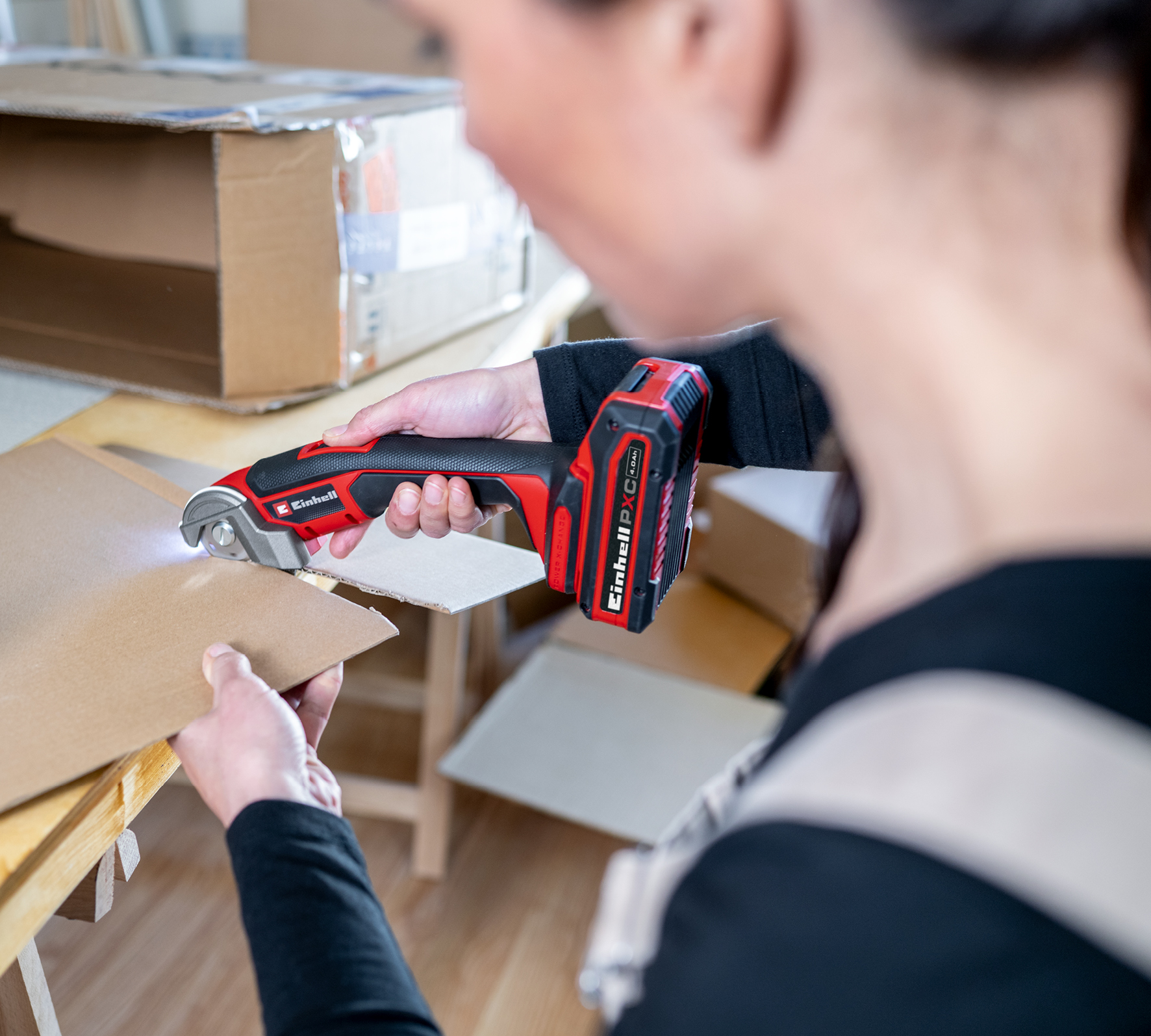 Eine Person verwendet einen rot-schwarzen Elektroschneider, um Pappe zu schneiden. Die Szene ist drinnen mit Kartons im Hintergrund.