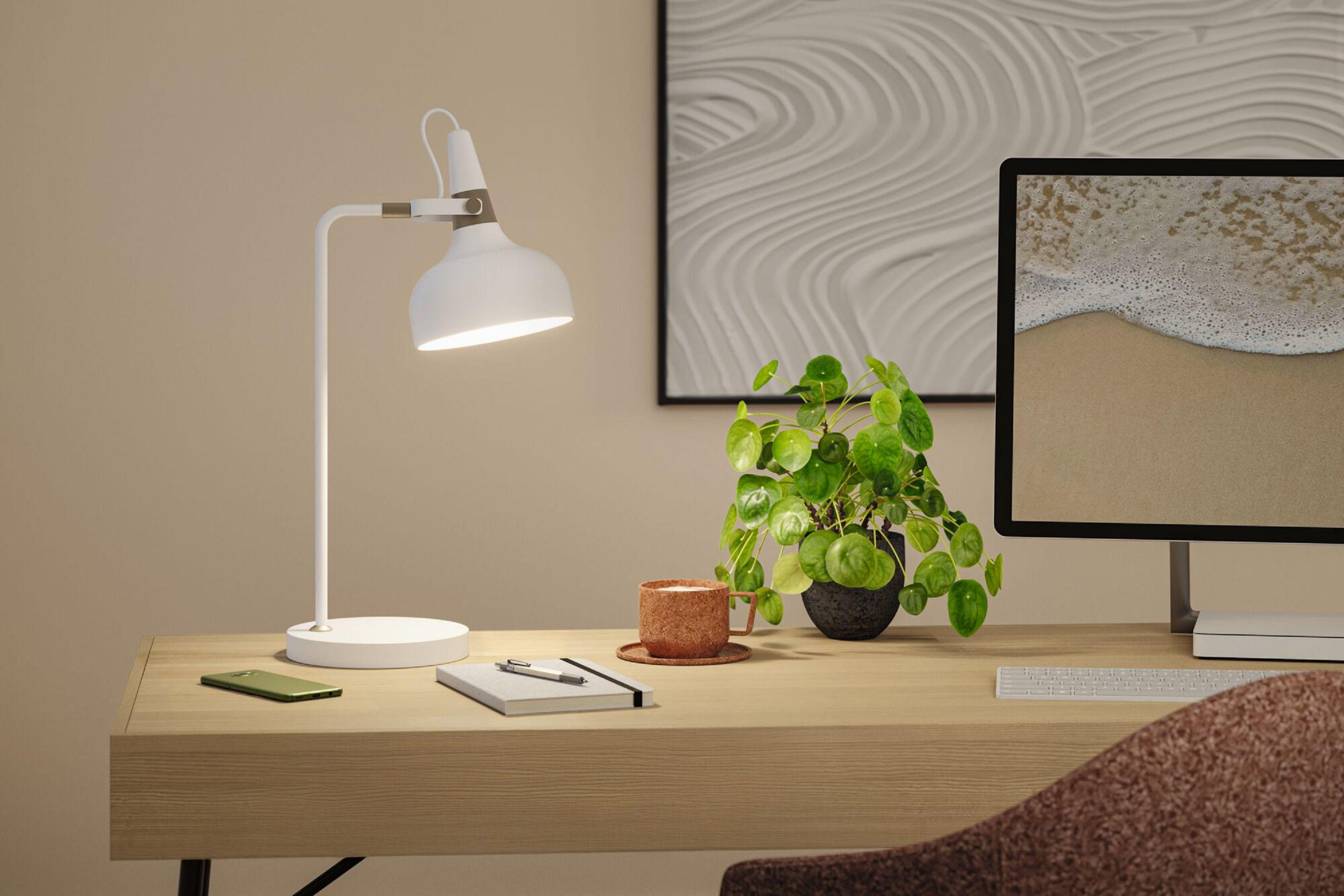 Biurko z lampą, rośliną, monitorem z plażą, kubkiem i notatnikiem.