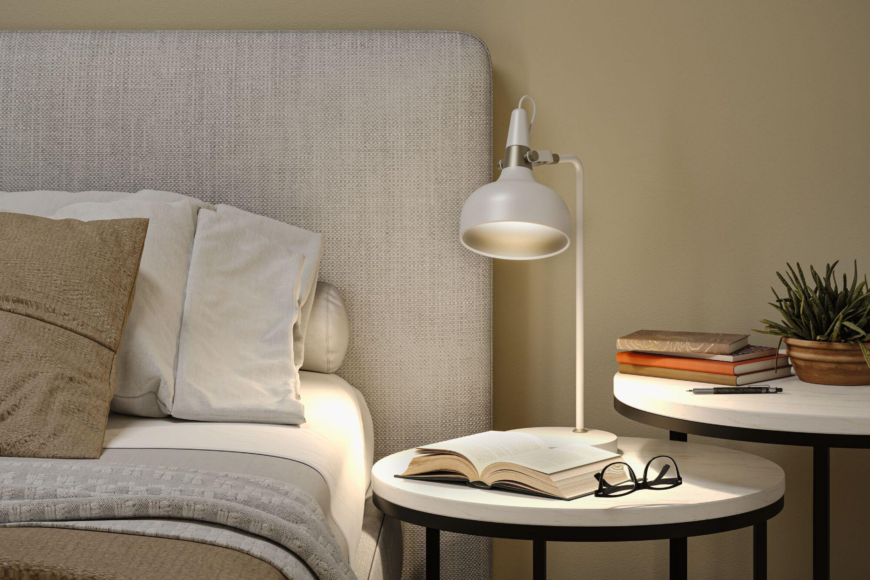 Scena sypialni z łóżkiem, stolikami nocnymi, lampą, książkami i okularami. Beżowe ściany i wezgłowie. Książki i roślina.