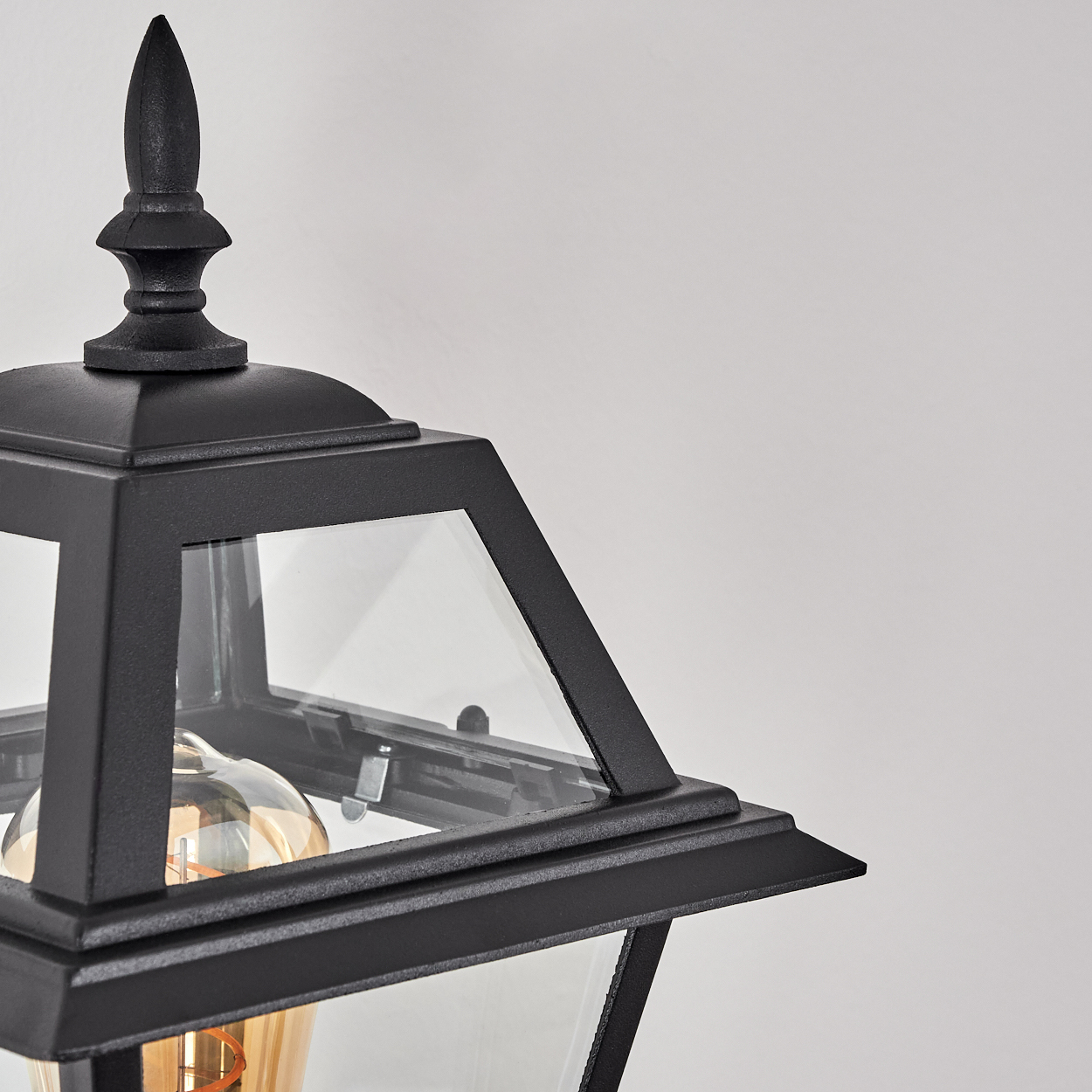 Nahaufnahme einer schwarzen Außenlampe mit Glasscheiben und einem dekorativen Aufsatz. Eine Glühbirne ist sichtbar. Weißer Hintergrund.