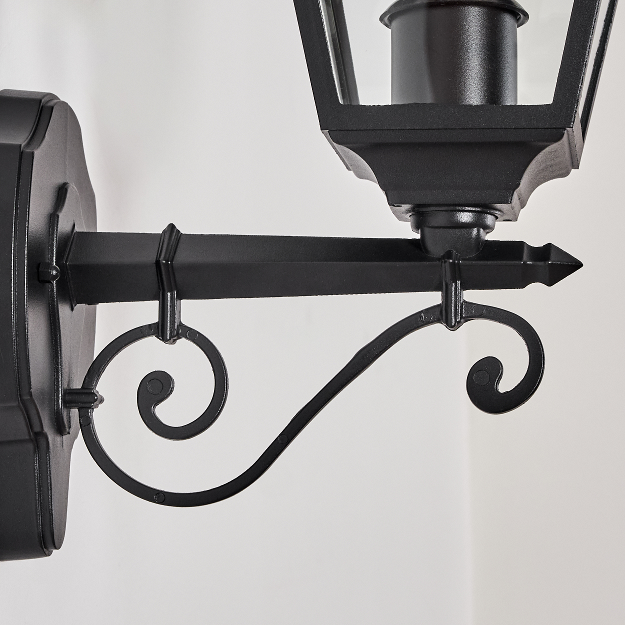 Eine schwarze Wandlaterne. Sie hat eine dekorative Halterung mit einem Schnörkeldesign. Der Hintergrund ist eine schlichte weiße Wand.