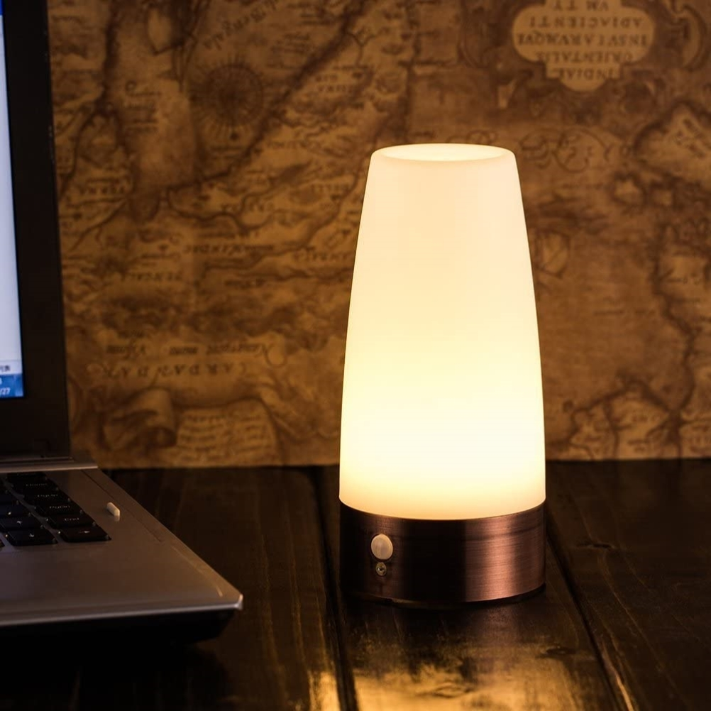 Eine leuchtende, weiße Lampe mit bronzefarbenem Sockel steht auf einem Holztisch neben einem Laptop. Im Hintergrund hängt eine Karte.