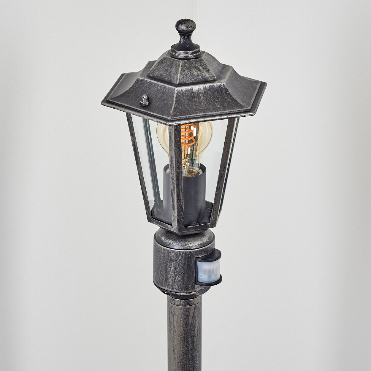 Dekorative Außenlaterne. Sie hat ein Glasgehäuse mit einer Glühbirne. Der Pfosten ist metallisch grau.