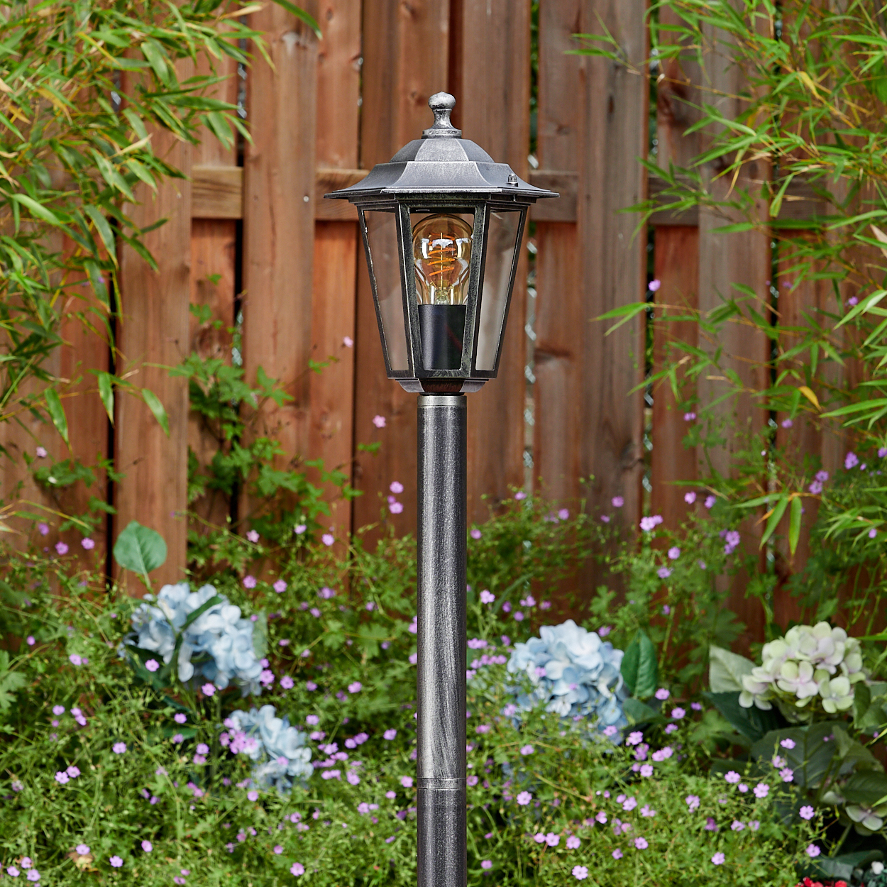 Eine dekorative Gartenlampe mit leuchtender Glühbirne steht vor einem Holzzaun, umgeben von Blumen und Grün.