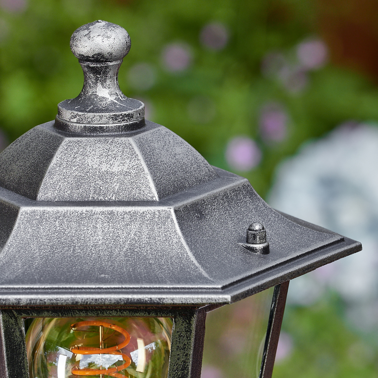 Nahaufnahme einer dekorativen Außenlampe mit silberner Oberfläche. Die Lampe hat eine Glühbirne mit Spiralfaden, vor unscharfem grünem Hintergrund.