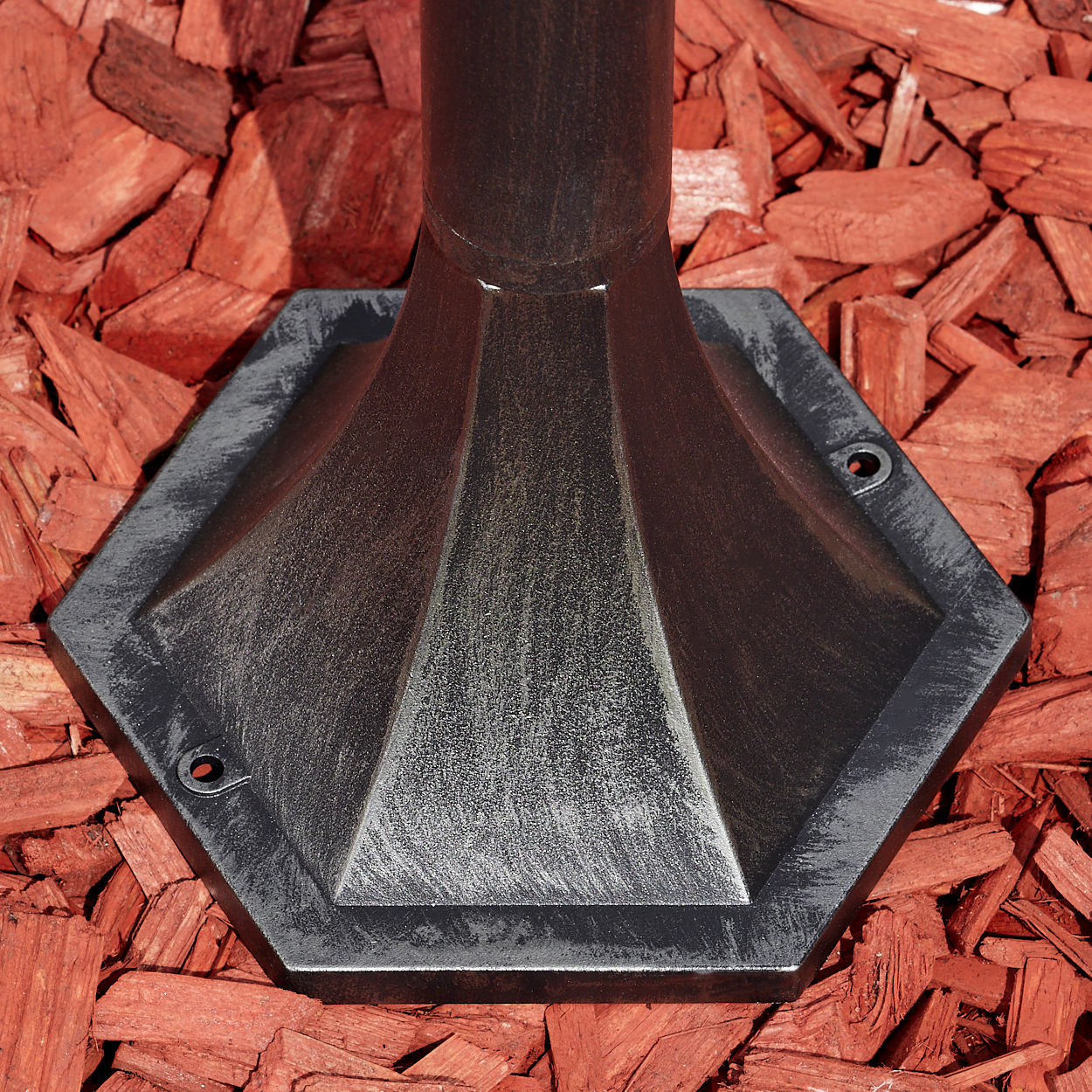 Dunkle Metallbasis einer Lampe auf roter Mulch. Die Basis ist sechseckig mit einem kegelförmigen Mittelteil.