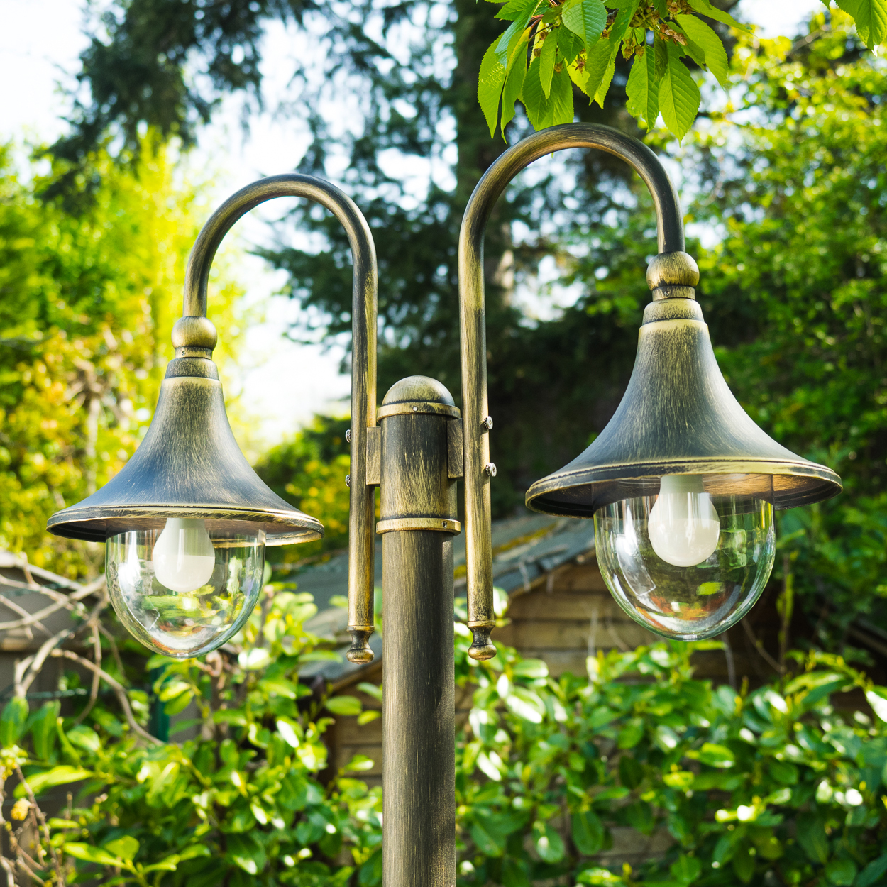 Eine verzierte Außenlampe mit zwei Lichtern, vor dem Hintergrund von grünem Laub und einem Holzschuppen.