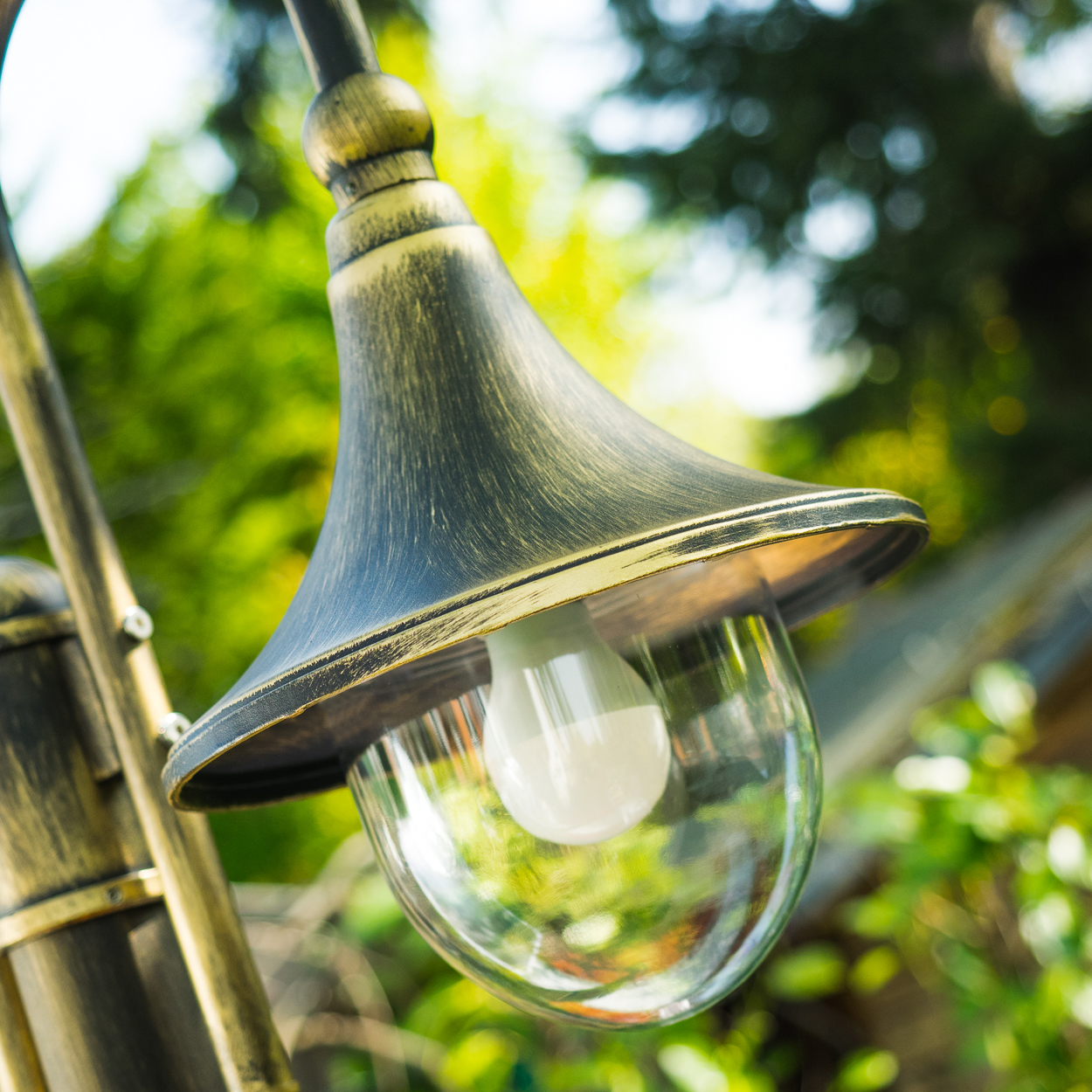 Nahaufnahme einer Vintage-Außenlampe mit Glasschirm und weißer Glühbirne. Die Lampe hat eine Bronzeausführung, vor grünem Laub.