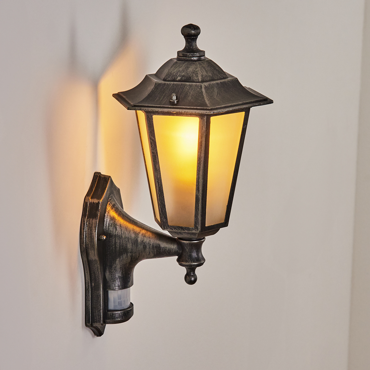 Eine Außenwandleuchte mit Bewegungssensor. Die Lampe ist schwarz mit goldenen Akzenten und das Licht ist an. Sie ist an einer weißen Wand montiert.