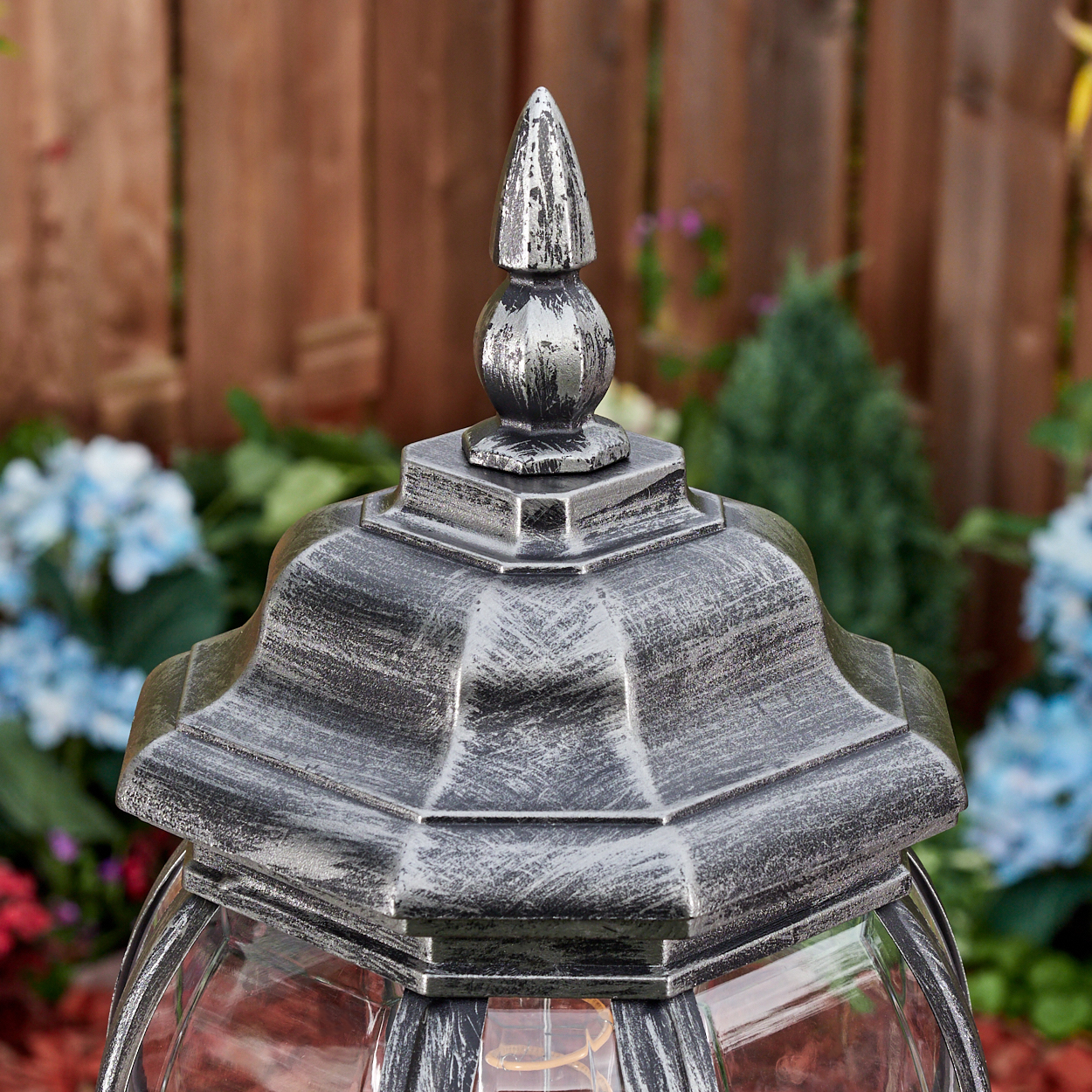 Nahaufnahme einer silbernen Außenlampe. Die Lampe hat eine dekorative Spitze mit einem spitzen Knauf. Blumen und ein Holzzaun sind im Hintergrund.