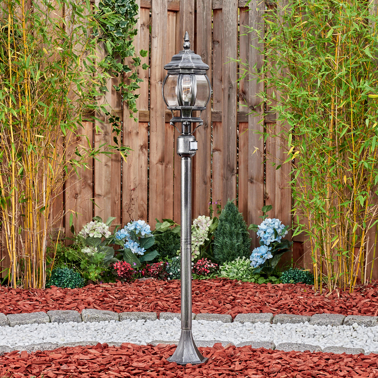 Eine dekorative Gartenlampe steht in einem Blumenbeet mit Bambus und einem Holzzaun im Hintergrund. Roter Mulch und weiße Steine vervollständigen die Szene.