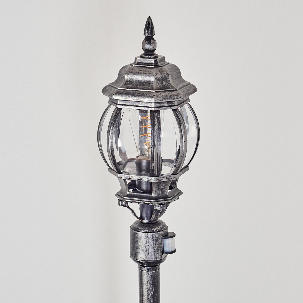 Eine dekorative silberne Außenlampe. Sie hat eine Glaskugel, eine spitze Spitze und einen Bewegungssensor. Der Hintergrund ist weiß.