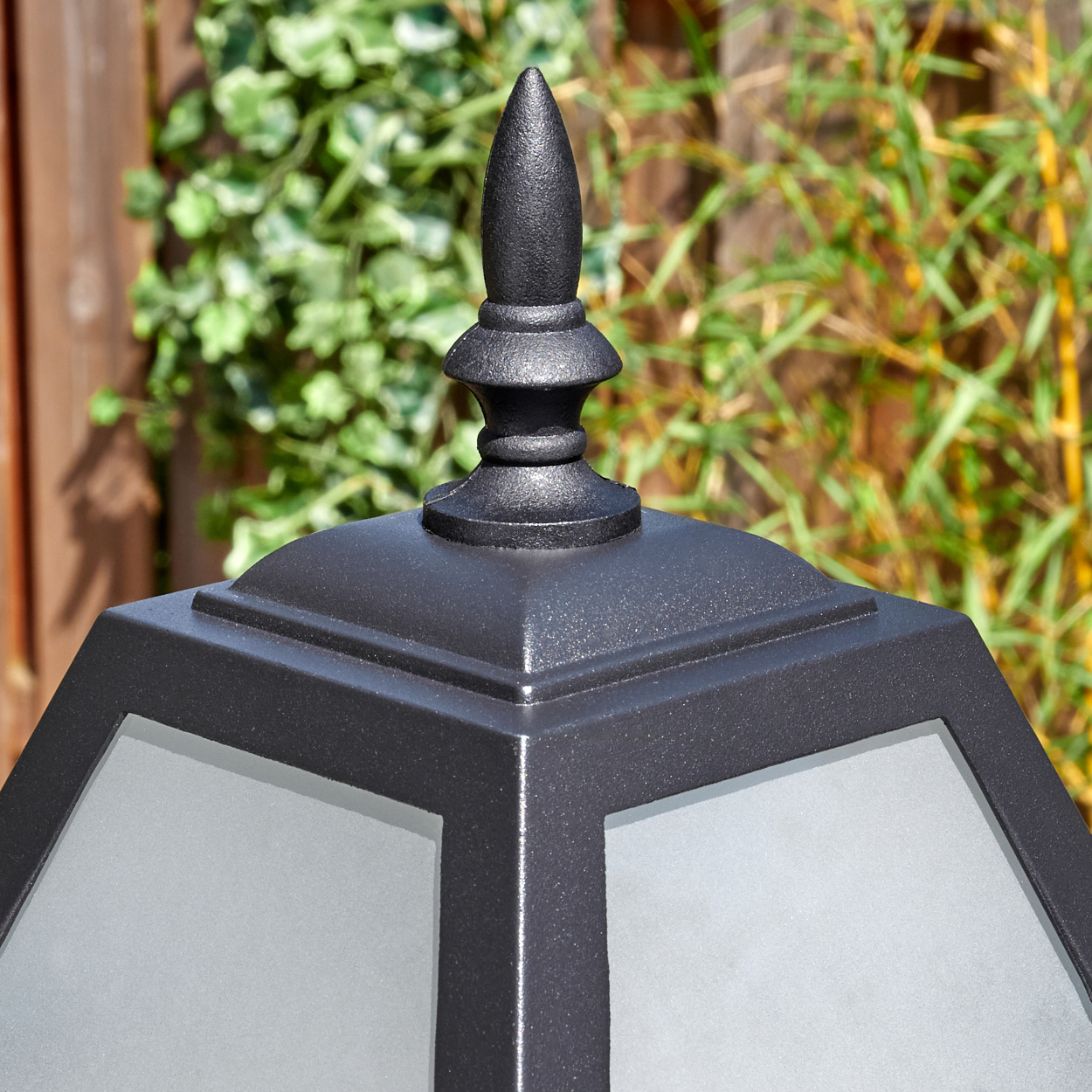 Nahaufnahme einer schwarzen Außenlampe mit spitzem Aufsatz. Grünes Laub und Bambus im Hintergrund. Links ein Holzzaun.