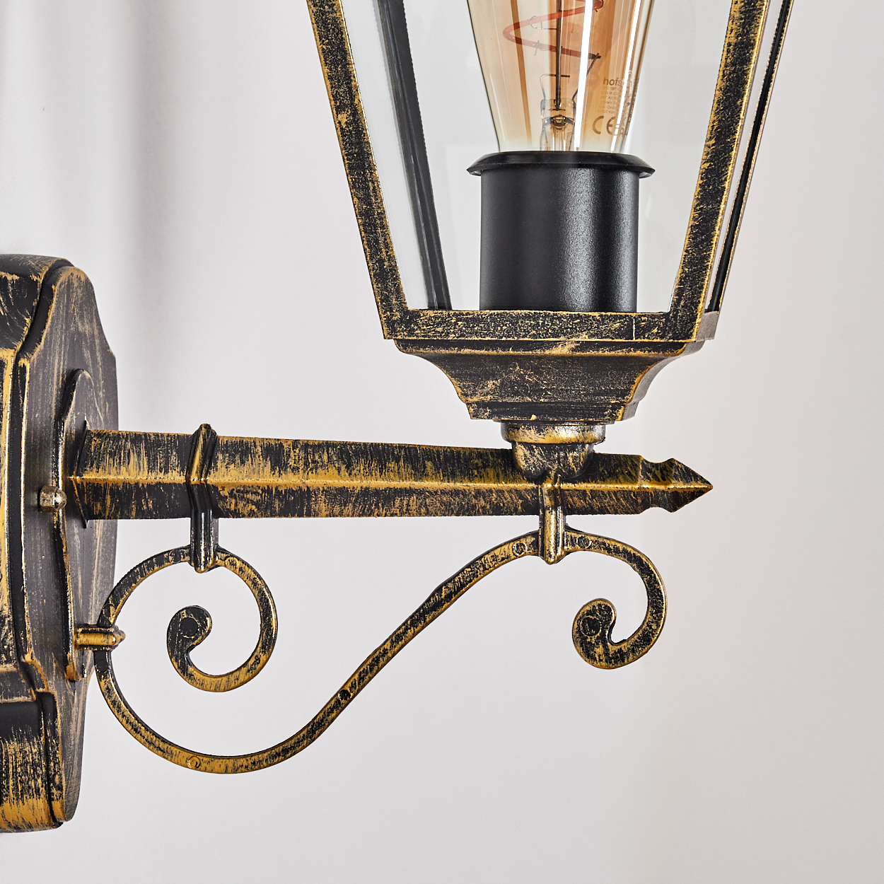 Nahaufnahme einer Wandlaterne. Die Lampe hat eine schwarze Glühbirnenfassung, Glasscheiben und einen dekorativen, goldfarbenen Metallrahmen.