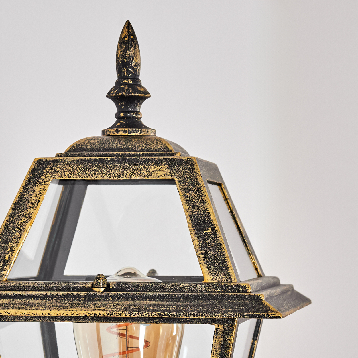 Nahaufnahme einer schwarz-goldenen Außenlampe. Sie hat Glasscheiben und einen dekorativen oberen Zierrat. Der Hintergrund ist weiß.