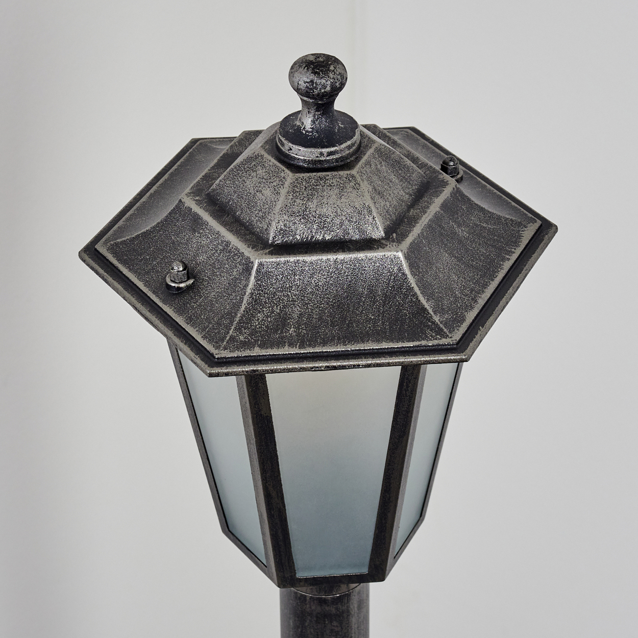 Eine dekorative Außenlampe. Sie hat eine sechseckige Oberseite und einen Glasteil zur Lichtausstrahlung. Die Lampe steht auf einem Pfahl vor weißem Hintergrund.