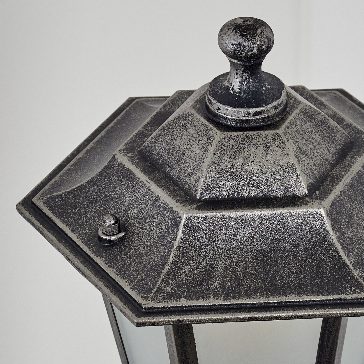 Nahaufnahme einer dekorativen Außenlampe. Sie hat eine metallische, gealterte Oberfläche mit sechseckigem Deckel und rundem Knauf. Der Hintergrund ist weiß.