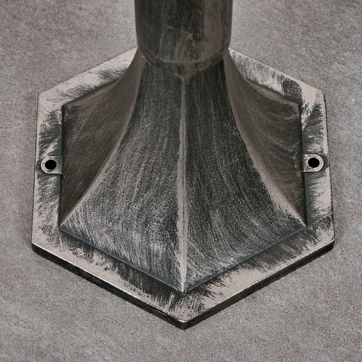 Nahaufnahme eines dekorativen Metallsockels. Sechseckige Form, strukturierte Silber- und Grautöne, mit zwei Schraubenlöchern, auf grauem Untergrund.