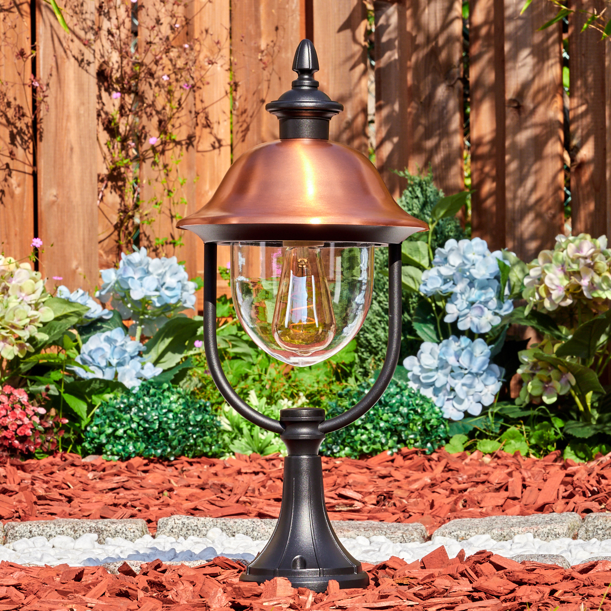 Eine dekorative Außenlampe mit kupferfarbener Oberseite und Glaskugel. Sie steht in einem Gartenbeet mit Mulch, Blumen und einem Holzzaun.