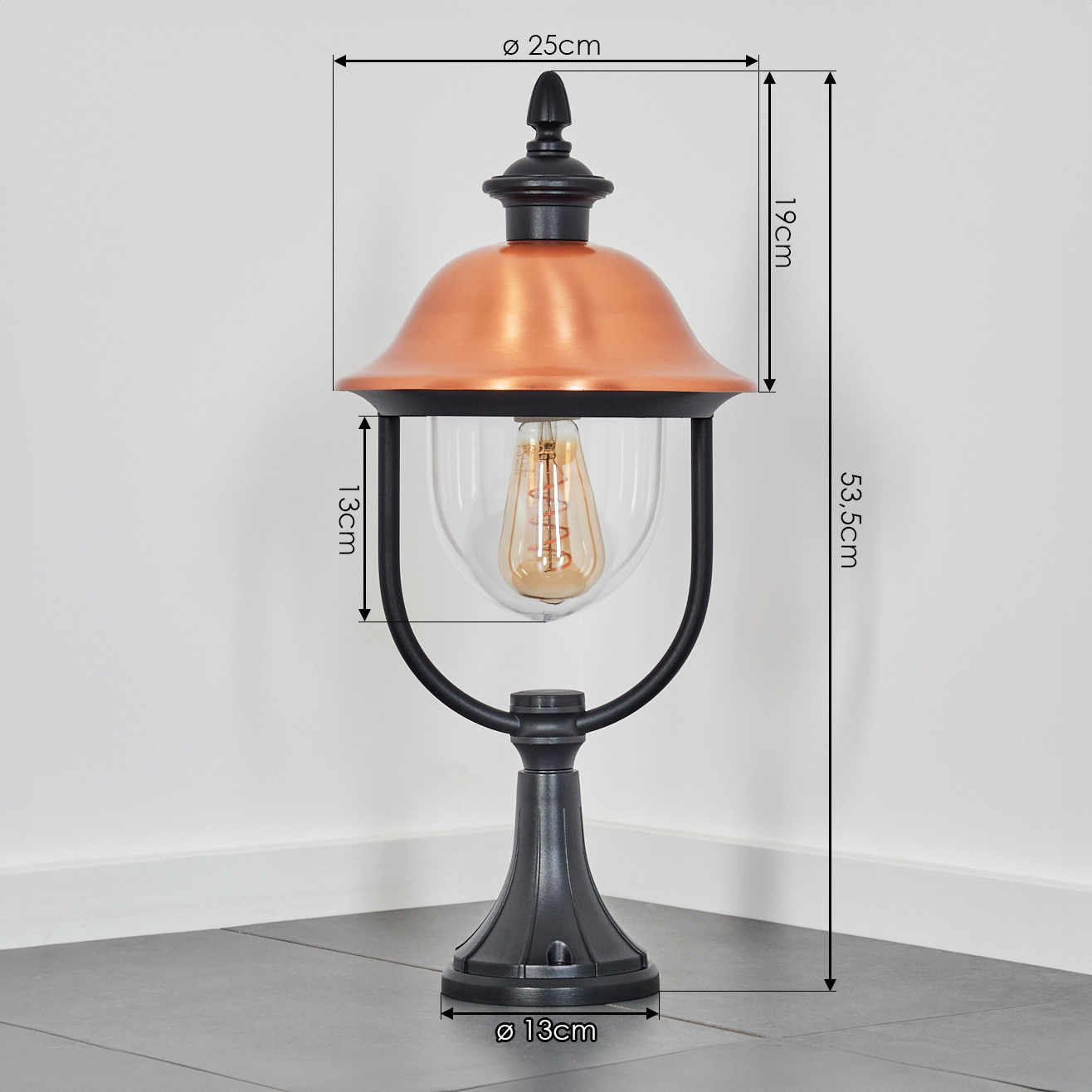 Eine dekorative Außenlampe. Sie hat einen schwarzen Sockel und Rahmen mit kupferfarbener Oberseite. Ein klares Glas umschließt eine Glühbirne.
