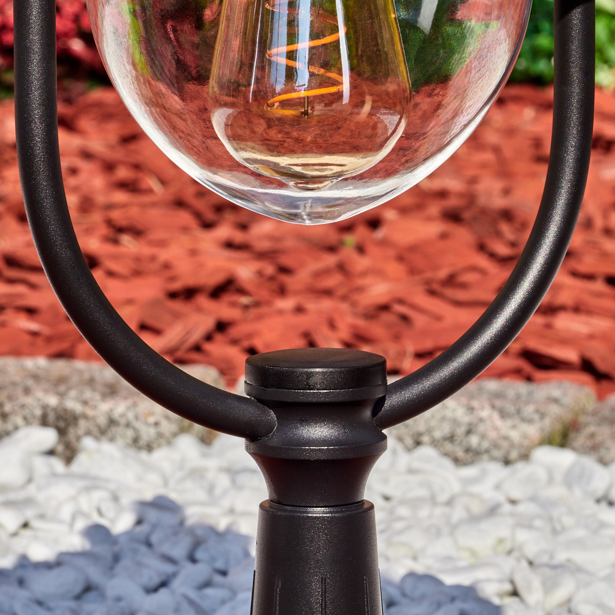 Nahaufnahme einer Lampe mit Glaskugel, schwarzem Metallrahmen und sichtbarer Glühbirne. Der Hintergrund hat roten Mulch und weiße Steine.