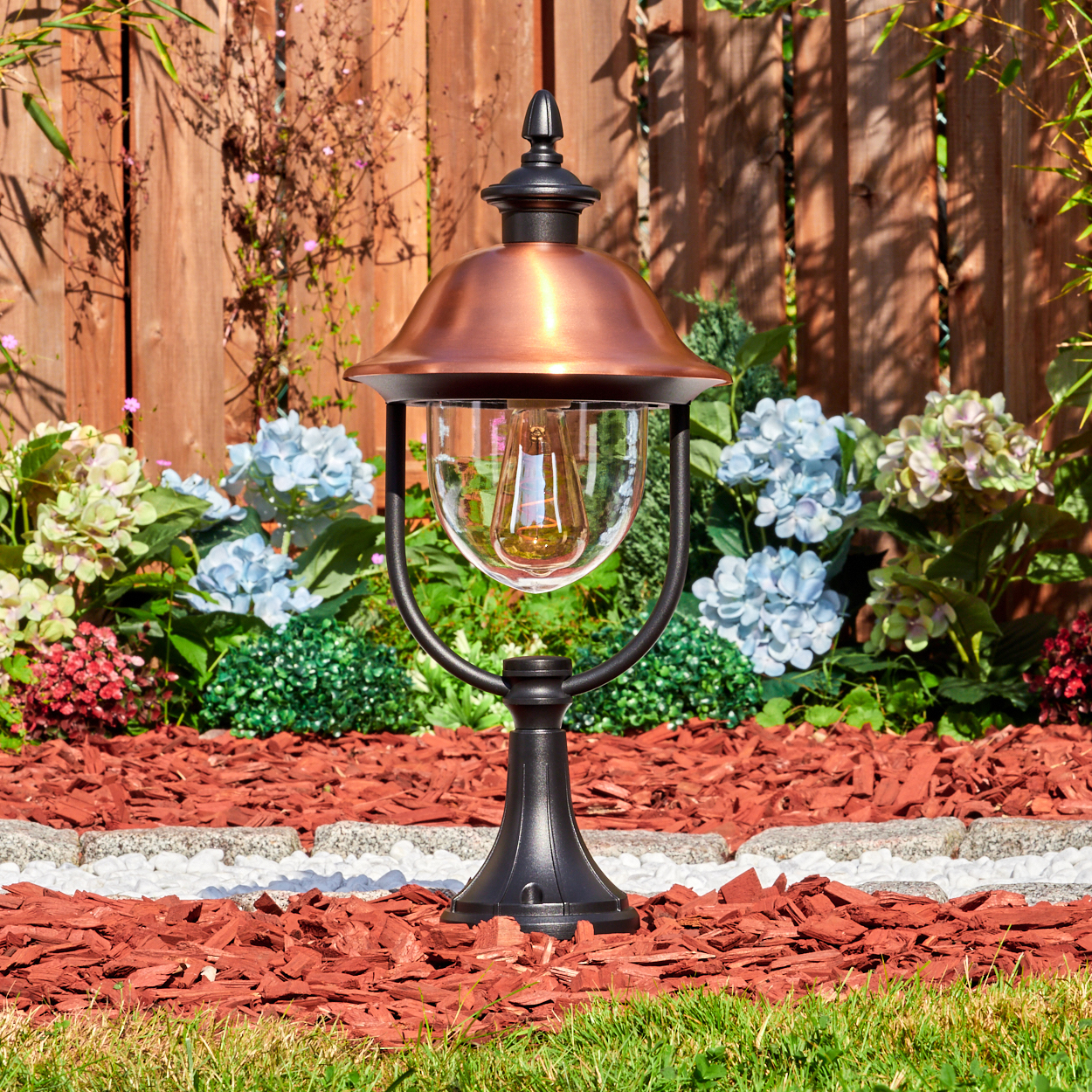 Eine dekorative Gartenlampe mit Kupferaufsatz und Glaskugel. Sie steht in einem Gartenbeet mit Blumen und Holzschnitzeln vor einem Holzzaun.
