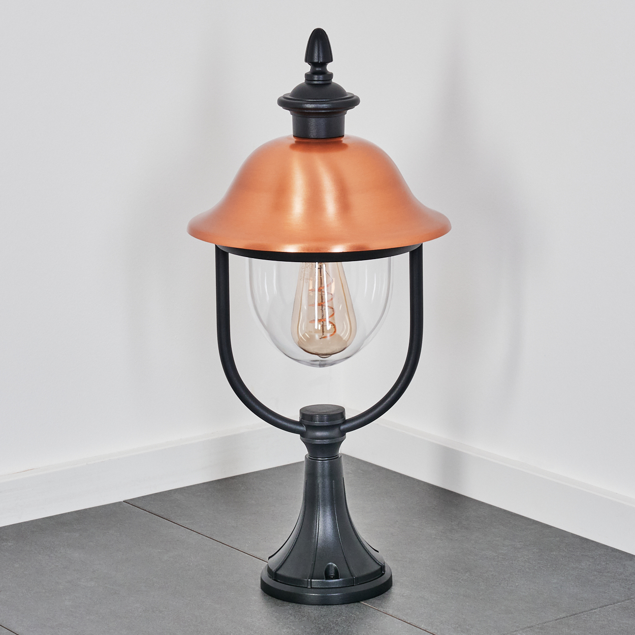 Eine dekorative Außenlampe mit kupferfarbenem Schirm, Glaskugel und schwarzem Sockel. Steht auf grau gefliestem Boden vor weißer Wand.