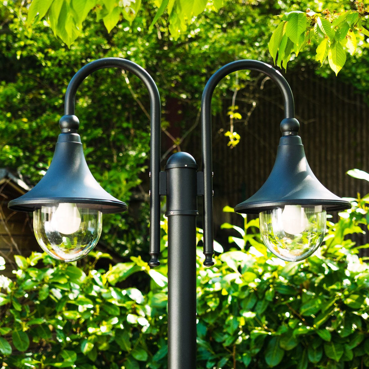 Eine schwarze Außenlampe mit zwei Lichtern. Grünes Laub im Hintergrund. Die Lampenschirme sind dunkelblau und die Glühbirnen sind sichtbar.