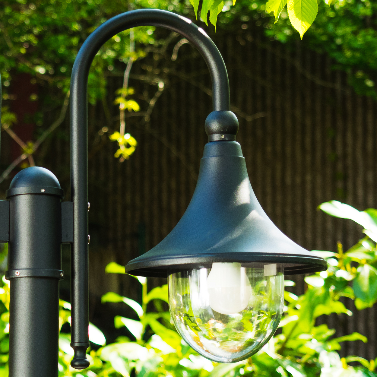 Eine schwarze Außenlampe. Sie hat einen gebogenen Arm und einen Glasschirm mit einer weißen Glühbirne. Grünes Laub ist im Hintergrund.