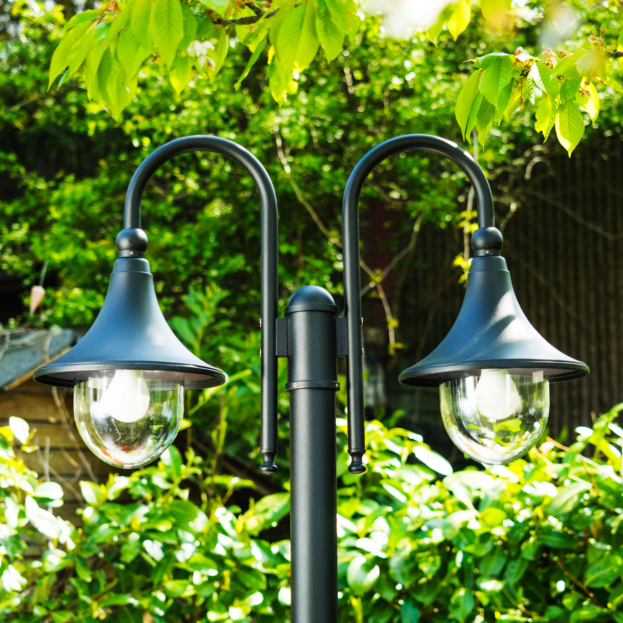 Eine schwarze Außenlampe mit zwei Lichtern, eingebettet in einen grünen Garten. Im Hintergrund sind die Blätter der Pflanzen und Bäume zu sehen.