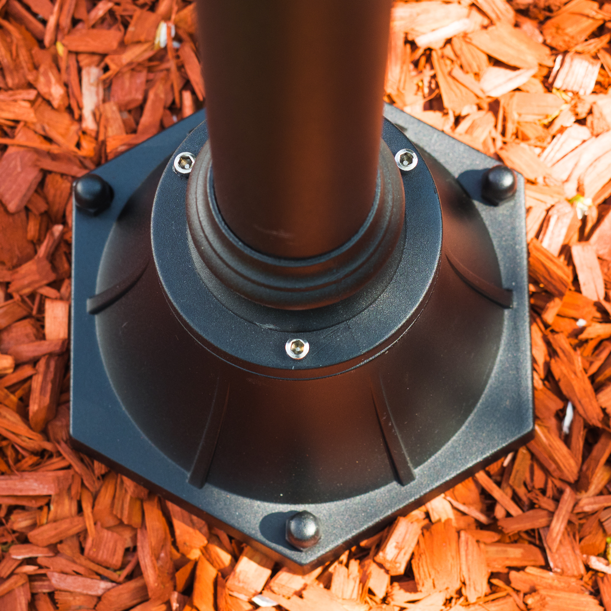 Schwarzer Metallsockel, sechseckig, mit Schrauben, auf rotbraunem Mulch.