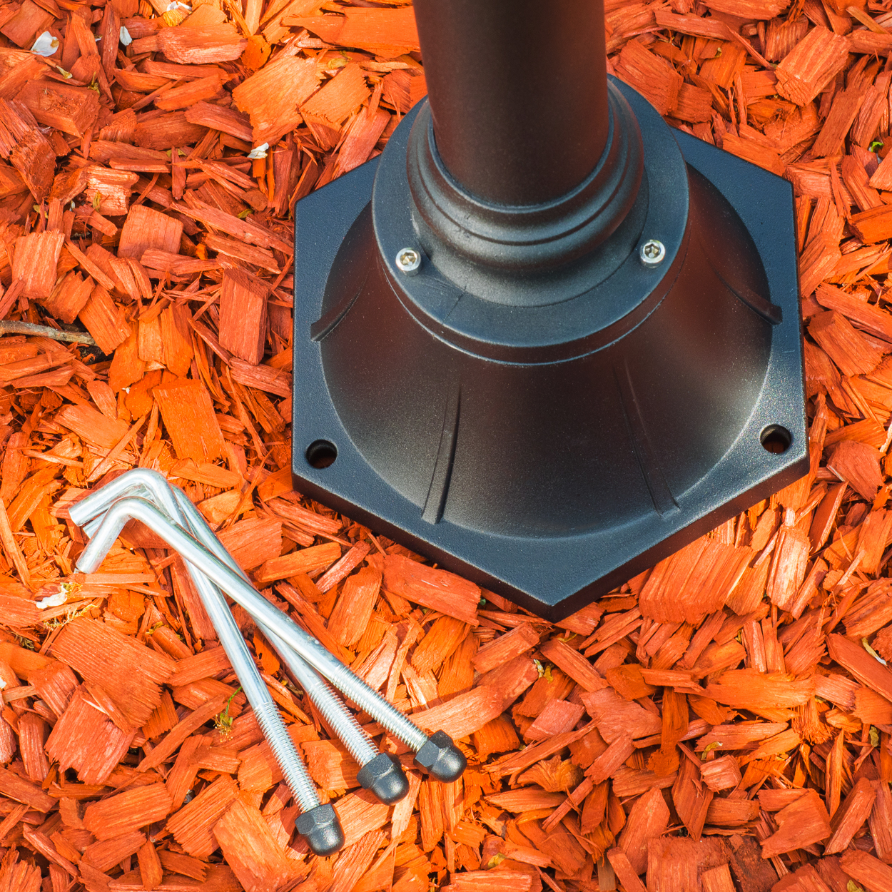 Schwarzer Lampensockel und -mast auf orangefarbenem Mulch, mit drei Metallankerschrauben. Die Basis ist sechseckig.