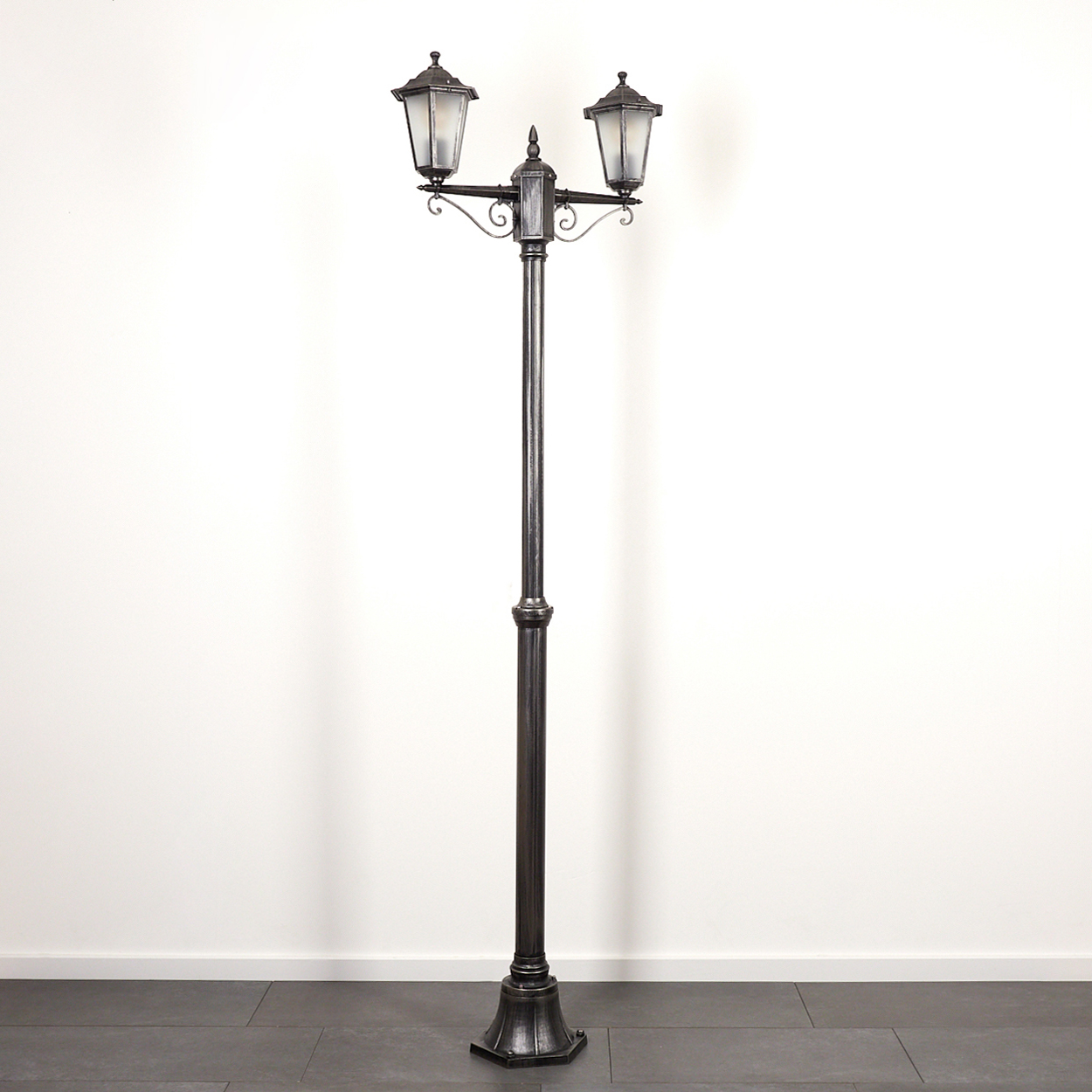 Eine dunkle Metalllaterne mit zwei Lampen. Sie steht auf einem gefliesten Boden vor einer weißen Wand.