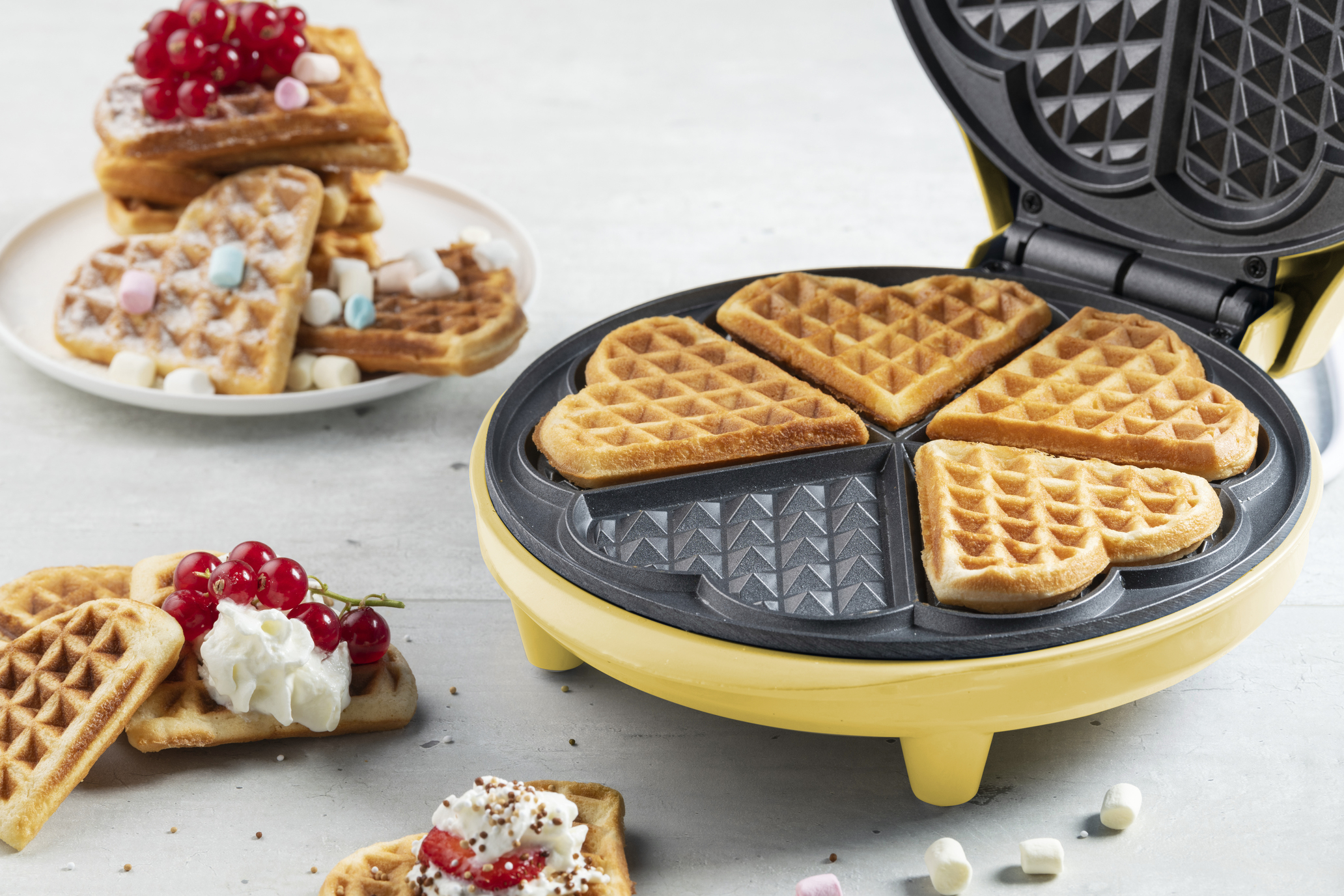 Gofr w kształcie serca w gofrownicy, z goframi na talerzu posypanymi czerwonymi jagodami i piankami.