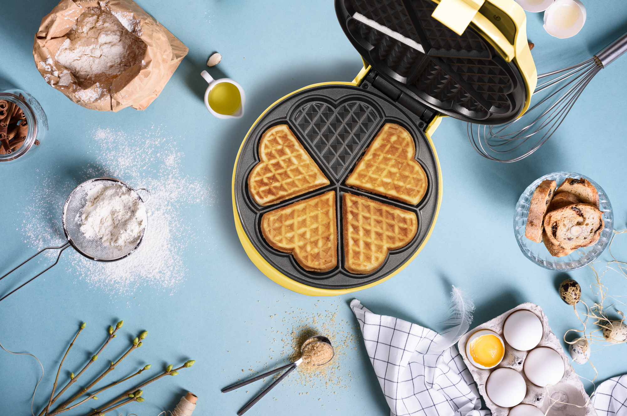 Widok z góry na gofrownicę z goframi w kształcie serca, otoczoną składnikami takimi jak mąka, jajka i cynamon na niebieskiej powierzchni.