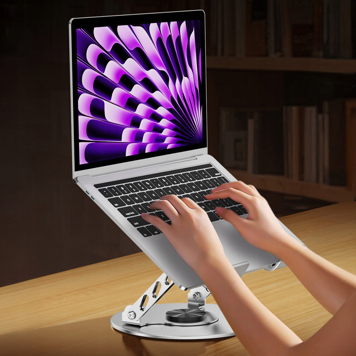 Laptop na srebrnym stojaku z piszącymi dłońmi. Ekran wyświetla fioletowy, abstrakcyjny wzór. Tło: drewniany stół i półka z książkami.