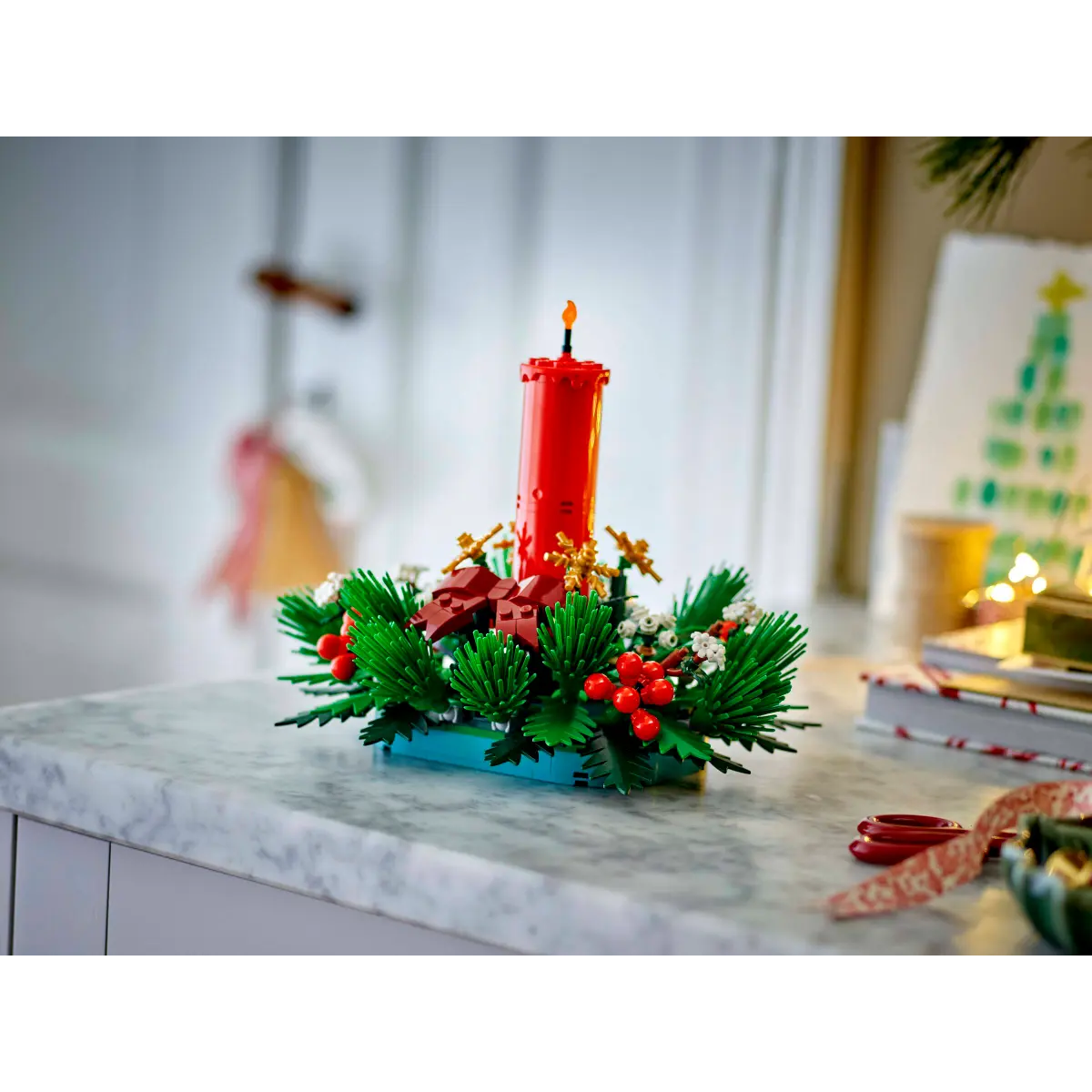 Bir Lego Noel dekorasyonu. Kırmızı bir mum, kırmızı meyveler, yeşil yapraklar ve altın detaylarla süslenmiş bir çelenk içinde beyaz bir masada duruyor.