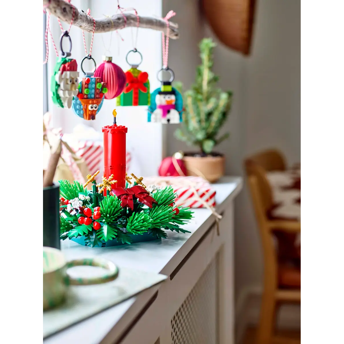 Asılı süslemelerle bir Noel sahnesi. Kırmızı bir mum ve yeşil bir çelenk beyaz bir yüzeyde duruyor. Arka planda küçük bir ağaç var.