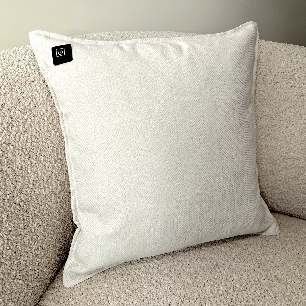Ein weißes Kissen mit einem Einschaltknopf liegt auf einer strukturierten beigefarbenen Couch. Das Kissen hat ein Fischgrätenmuster. Der Hintergrund ist eine Wand.
