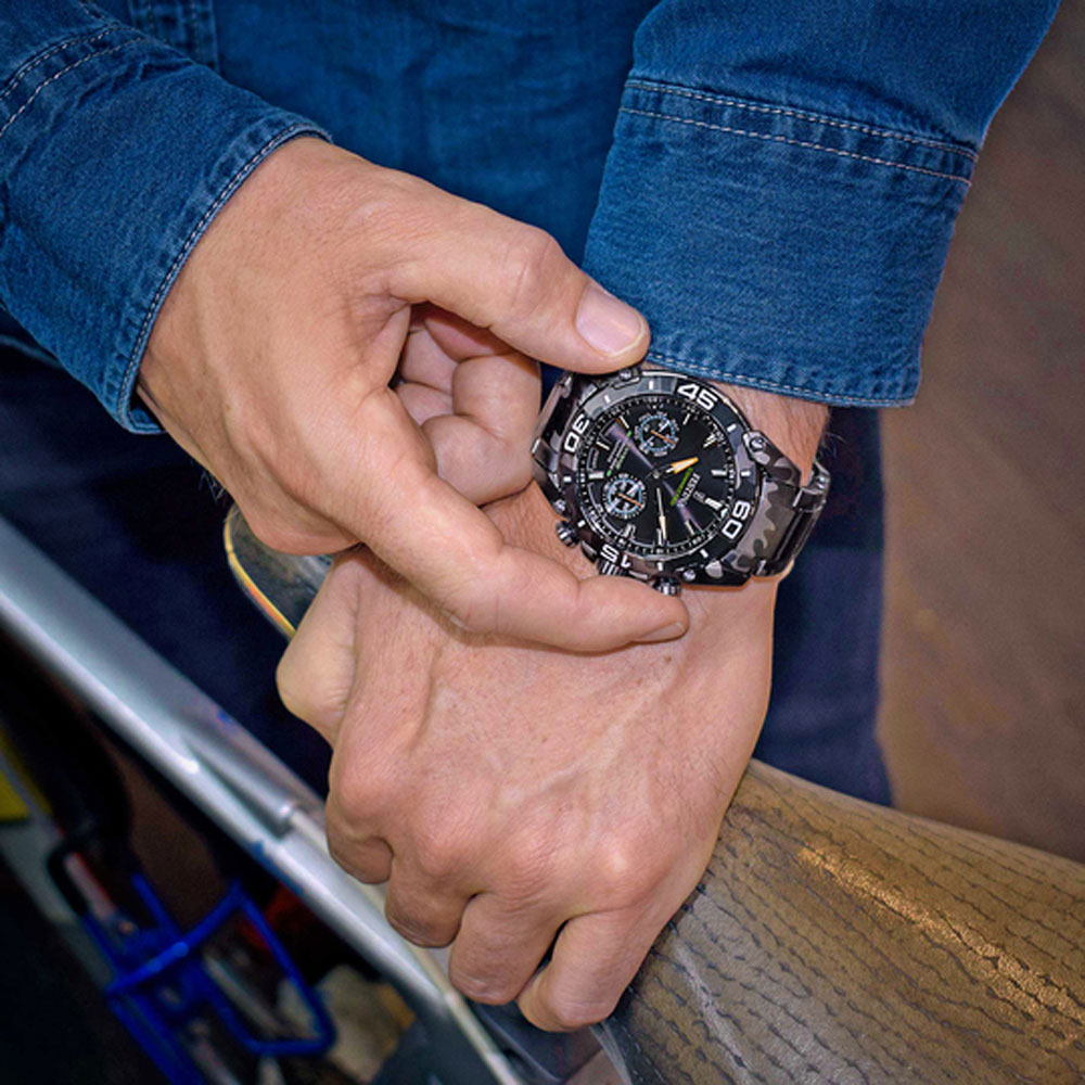 Eine Person in einem Jeanshemd hält eine schwarze Uhr mit Camouflage-Armband. Die Uhr hat ein schwarzes Zifferblatt mit weißen Markierungen.