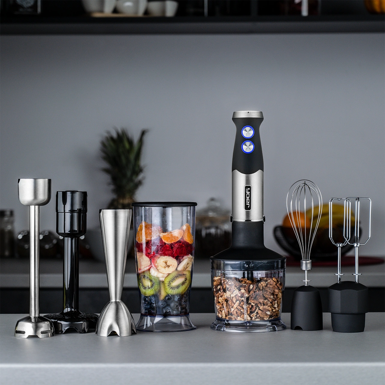 Blender z załącznikami, szklanka pełna owoców i orzechów włoskich. W tle kuchnia z ananasem i miskami.