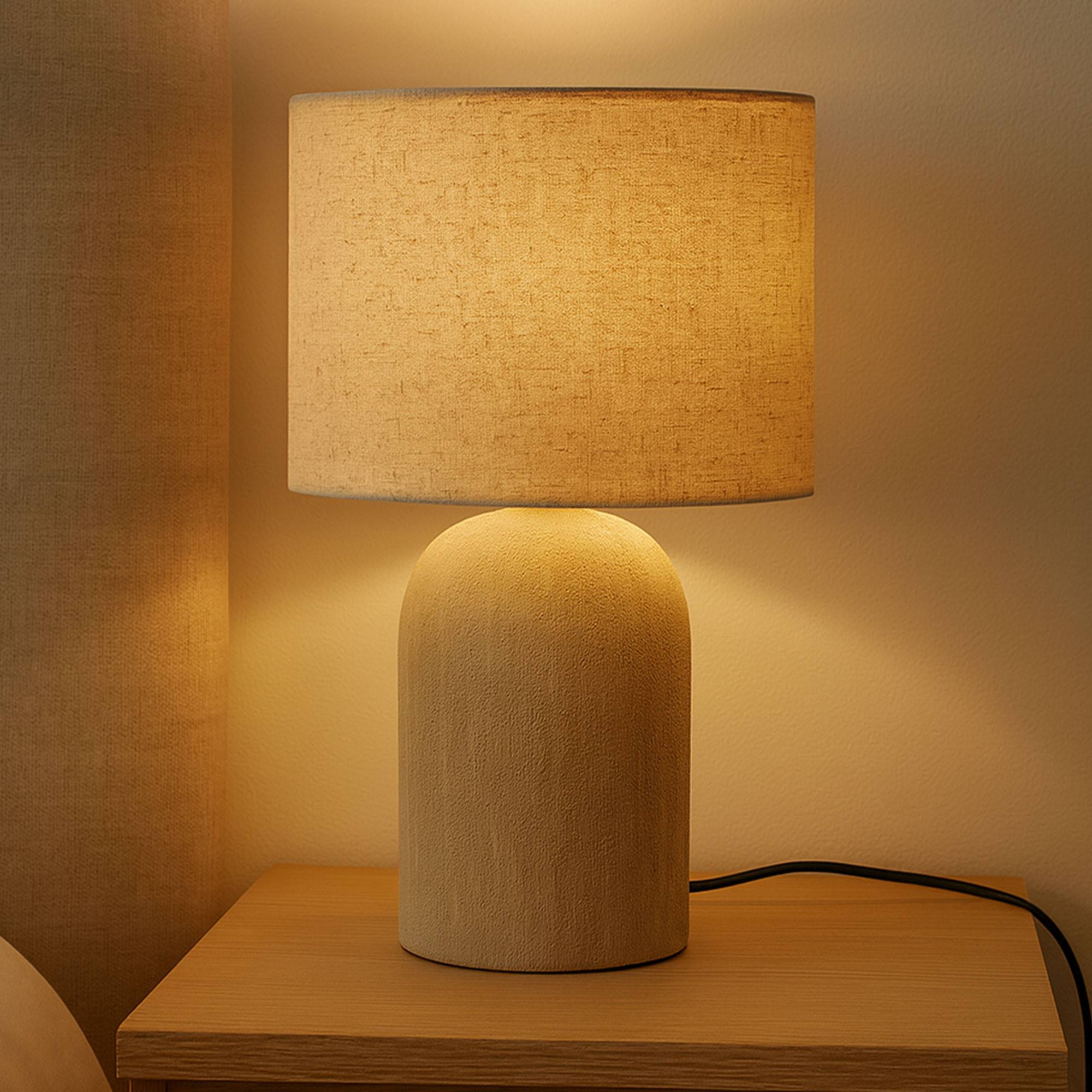 Una lámpara de mesa beige sobre una mesita de noche de madera. La lámpara tiene una pantalla cilíndrica y una base redondeada. Luz cálida.