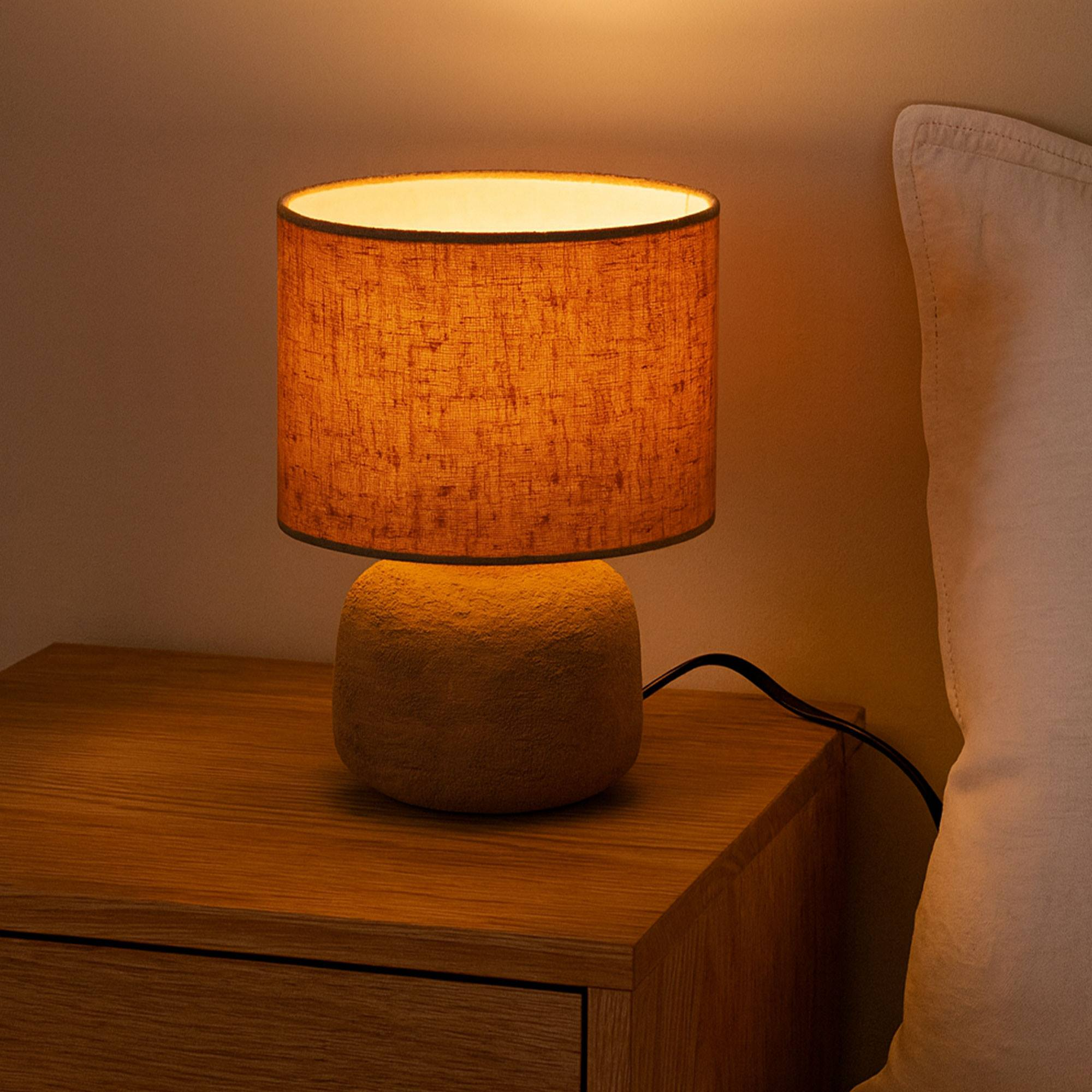 Una lámpara encendida está sobre una mesita de noche junto a una cama. La lámpara tiene una pantalla cilíndrica y una base redonda.