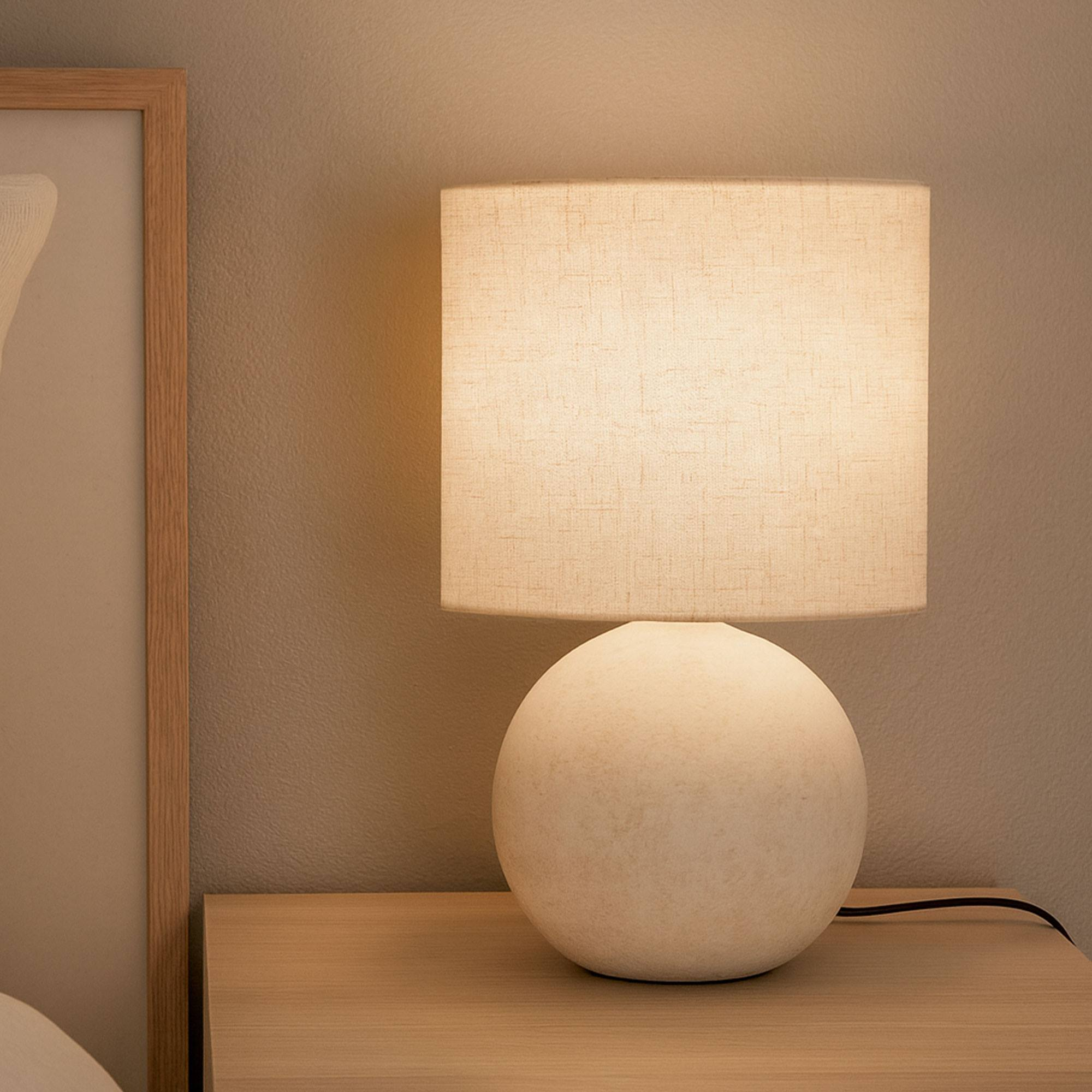 Una lámpara encendida con pantalla beige y base esférica blanca sobre superficie de madera. A la izquierda, un cuadro enmarcado.