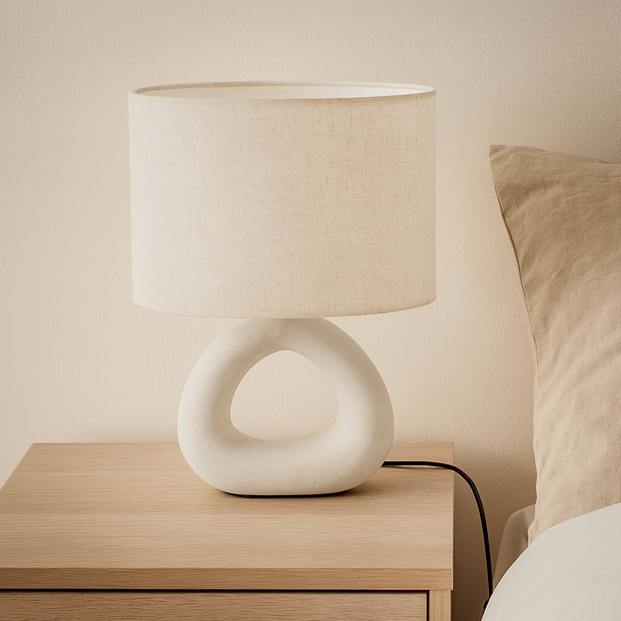 Una lámpara blanca con base circular sobre una mesita de noche de madera. Una almohada beige al fondo.