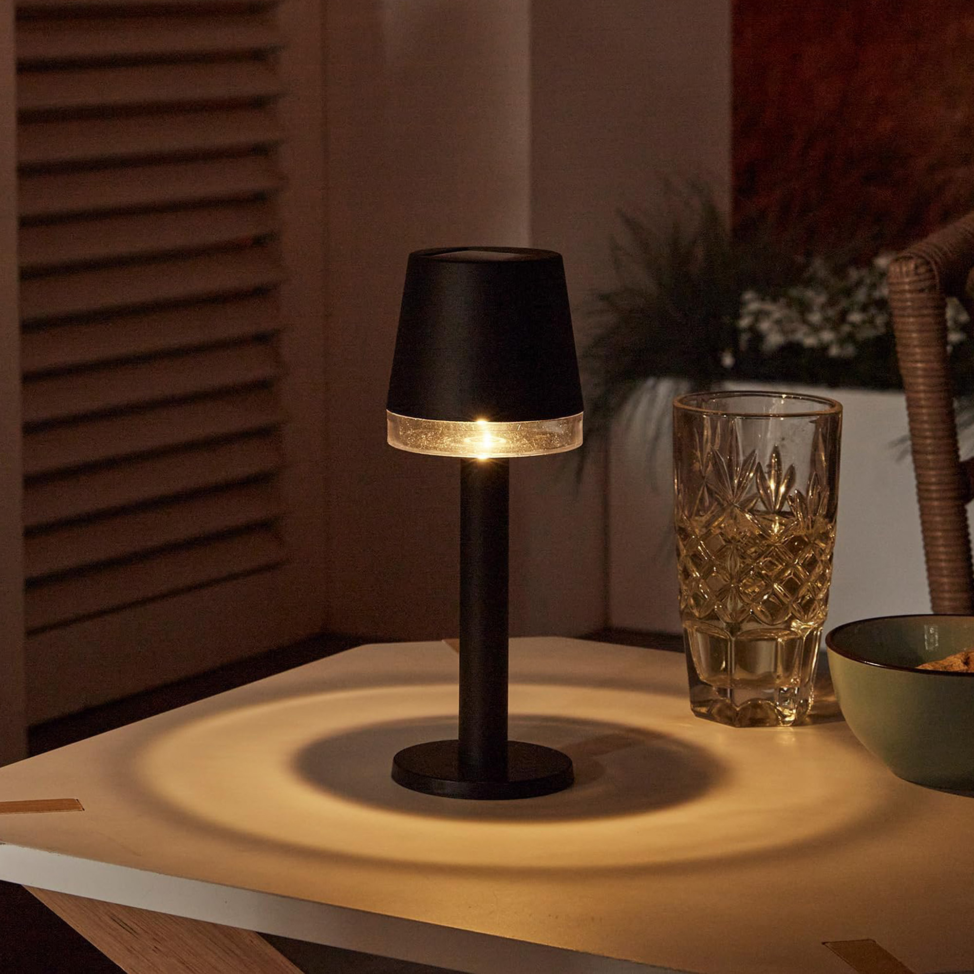 Una lámpara negra encendida sobre una mesa, junto a un vaso y un cuenco. La lámpara proyecta un círculo de luz.